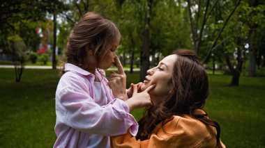 side view of happy mother touching nose of cute daughter in green park clipart