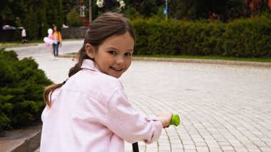 Pembe kot ceketli mutlu çocuk parkta scooter sürüyor. 