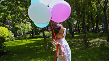 Çocuk renkli balonlar tutuyor ve parkta barış işareti gösteriyor.