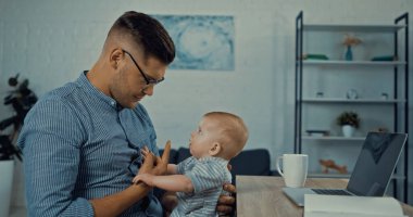 side view of father in glasses looking at infant boy while working from home 