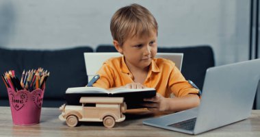 boy holding notebook and looking at laptop near wooden toy car on desk