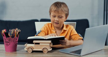 boy looking at notebook near laptop and wooden toy car on desk