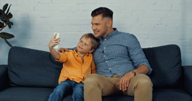 joyful father and cheerful son smiling while taking selfie at home 