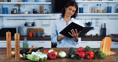 smiling housewife reading cookbook near raw vegetables and eggs in kitchen