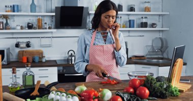 housewife with knife licking cut finger near chopped bell pepper and fresh vegetables