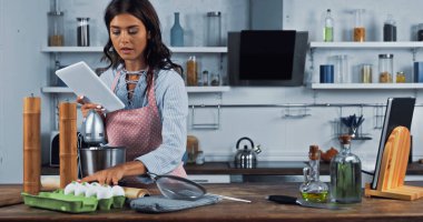 woman with digital tablet reading recipe near fresh eggs while cooking in kitchen