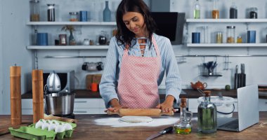 happy housewife in apron rolling out dough near food processor and laptop on worktop