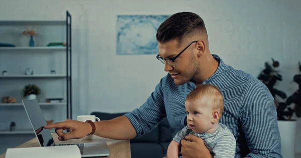 Joyful Freelancer Man Glasses Pointing Laptop While Holding Infant Boy — Stockfoto