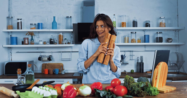 cheerful brunette woman with spice mills near raw vegetables and eggs in kitchen