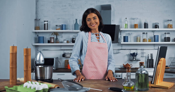 happy woman in apron near food processor and ingredients on kitchen worktop