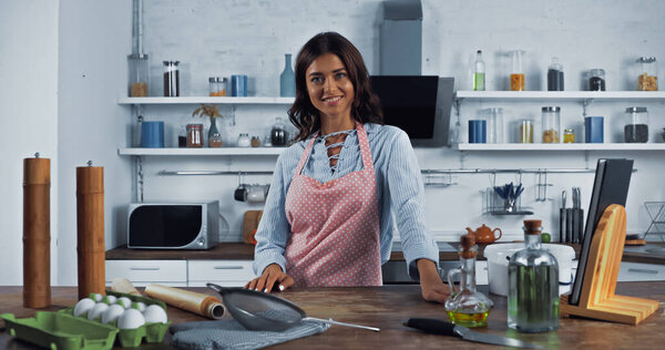 brunette housewife in apron smiling at camera near kitchenware and food on worktop