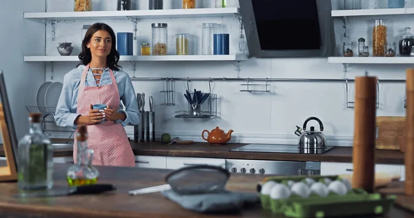 brunette woman with cup of tea smiling in kitchen near blurred worktop