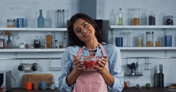 vegetarian woman in apron holding bowl of vegetable salad and smiling at camera