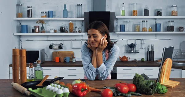 brunette woman smiling near fresh vegetables and cooking utensils on kitchen worktop