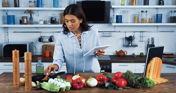 brunette woman with digital tablet reaching raw eggs near fresh vegetables on worktop