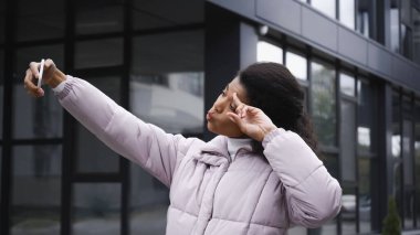curly african american woman in puffer jacket taking selfie on smartphone and showing peace sign outside