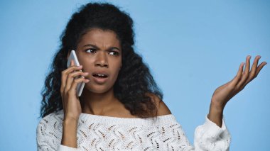 displeased african american woman in knitted sweater gesturing while talking on cellphone isolated on blue