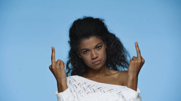 rude african american woman in white knitted sweater showing middle fingers isolated on blue