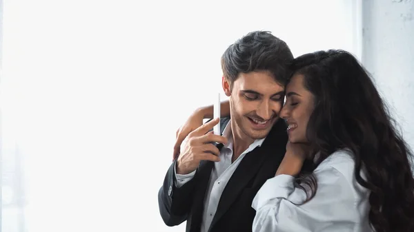Happy Brunette Woman Hugging Cheerful Boyfriend Suit Holding Smartphone — Stockfoto