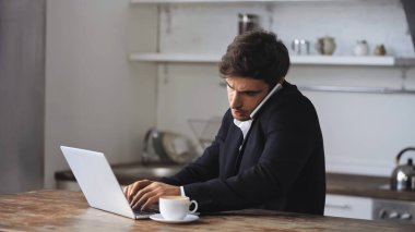 freelancer talking on smartphone near cup of coffee while using laptop 