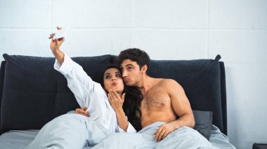 young woman sending air kiss while taking selfie with shirtless man in bed