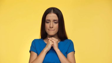 pensive young brunette woman in blue t-shirt standing with clenched hands isolated on yellow clipart