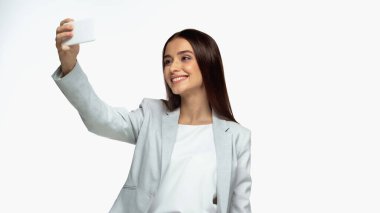 happy businesswoman in grey blazer taking selfie on smartphone isolated on white