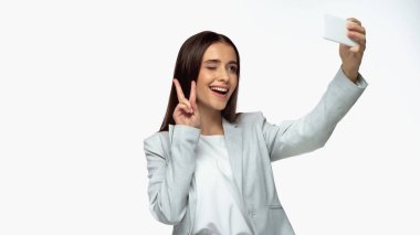 cheerful businesswoman in grey blazer showing peace sign and taking selfie isolated on white