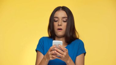 Mavi tişörtlü genç esmer kadın sarıya izole edilmiş akıllı telefonla yazarken yüzünü buruşturuyor.