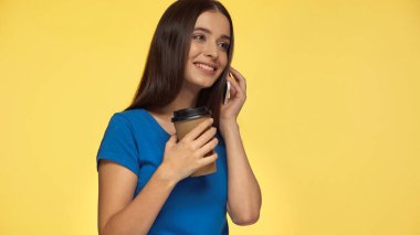 Mavi tişörtlü mutlu genç kadın akıllı telefonla konuşuyor ve sarı bardakta izole edilmiş kağıt bardak tutuyor.