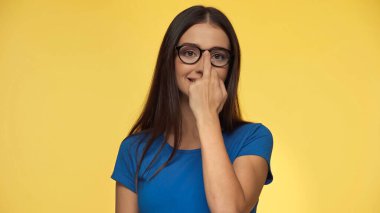 young brunette woman in blue t-shirt adjusting eyeglasses while showing middle finger isolated on yellow