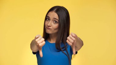young brunette woman in blue t-shirt showing dislike and biting lip isolated on yellow