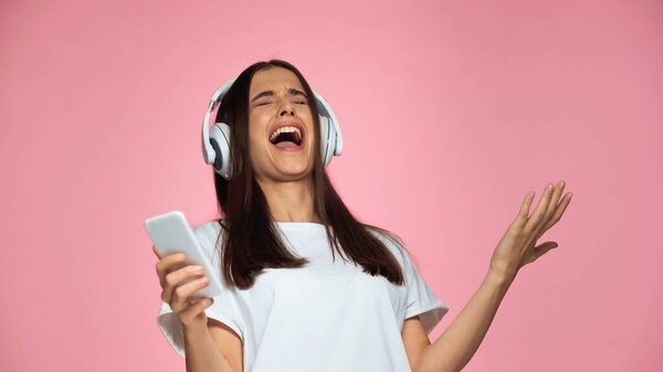 brunette young woman in wireless headphones singing and holding smartphone isolated on pink