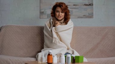 sick and redhead woman talking at camera while sitting near medication on coffee table 