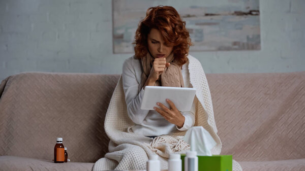 redhead sick woman using digital tablet and coughing near medication while sitting on couch