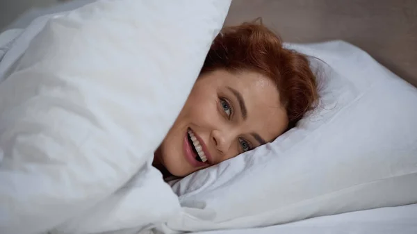 cheerful and redhead woman lying on pillow under white blanket