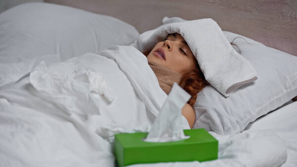 redhead woman sneezing in napkin while lying with towel on head under blanket 