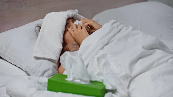 sick woman sneezing in napkin while lying with towel on head under blanket
