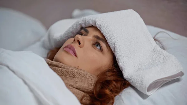 sick woman in scarf and with towel on head lying under blanket on bed