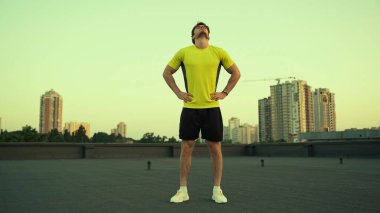 Young sportsman holding hands on hips while warming up on roof in evening 