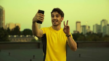Smiling sportsman taking selfie on smartphone and gesturing on roof in evening 