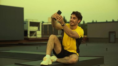 Smiling sportsman in sportswear and sneakers taking selfie on roof in evening 