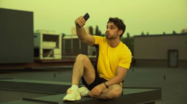 Brunette sportsman grimacing while taking selfie on roof in evening 