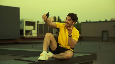 Cheerful sportsman taking selfie and showing secret gesture on roof in evening 