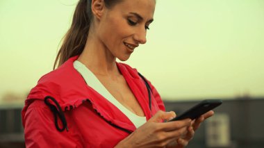 Positive brunette sportswoman using mobile phone on blurred roof in evening 