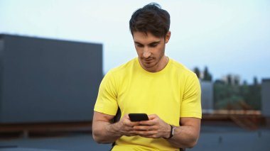 Sportsman in yellow sportswear using mobile phone on blurred roof in evening 