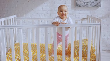 happy infant child standing in baby crib and smiling near brick wall