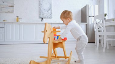 adorable toddler child playing with plastic building blocks on rocking horse in spacious apartment