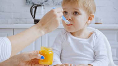 mother holding spoon and feeding baby boy with carrot puree in feeding chair 