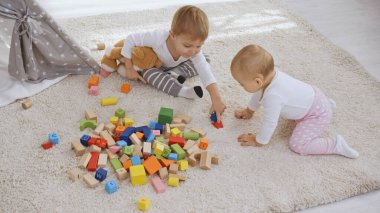 toddler boy sitting on carpet with teddy bear near baby sister and playing with wooden blocks together